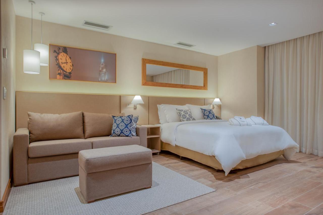 Wyndham Gramado Termas Resort & Spa Exterior foto The photo shows a modern hotel room featuring a cozy, well-decorated interior. There’s a large bed with white bedding, complemented by decorative pillows. Next to the bed is a stylish nightstand with a lamp. A comfortable sofa is positioned in one co