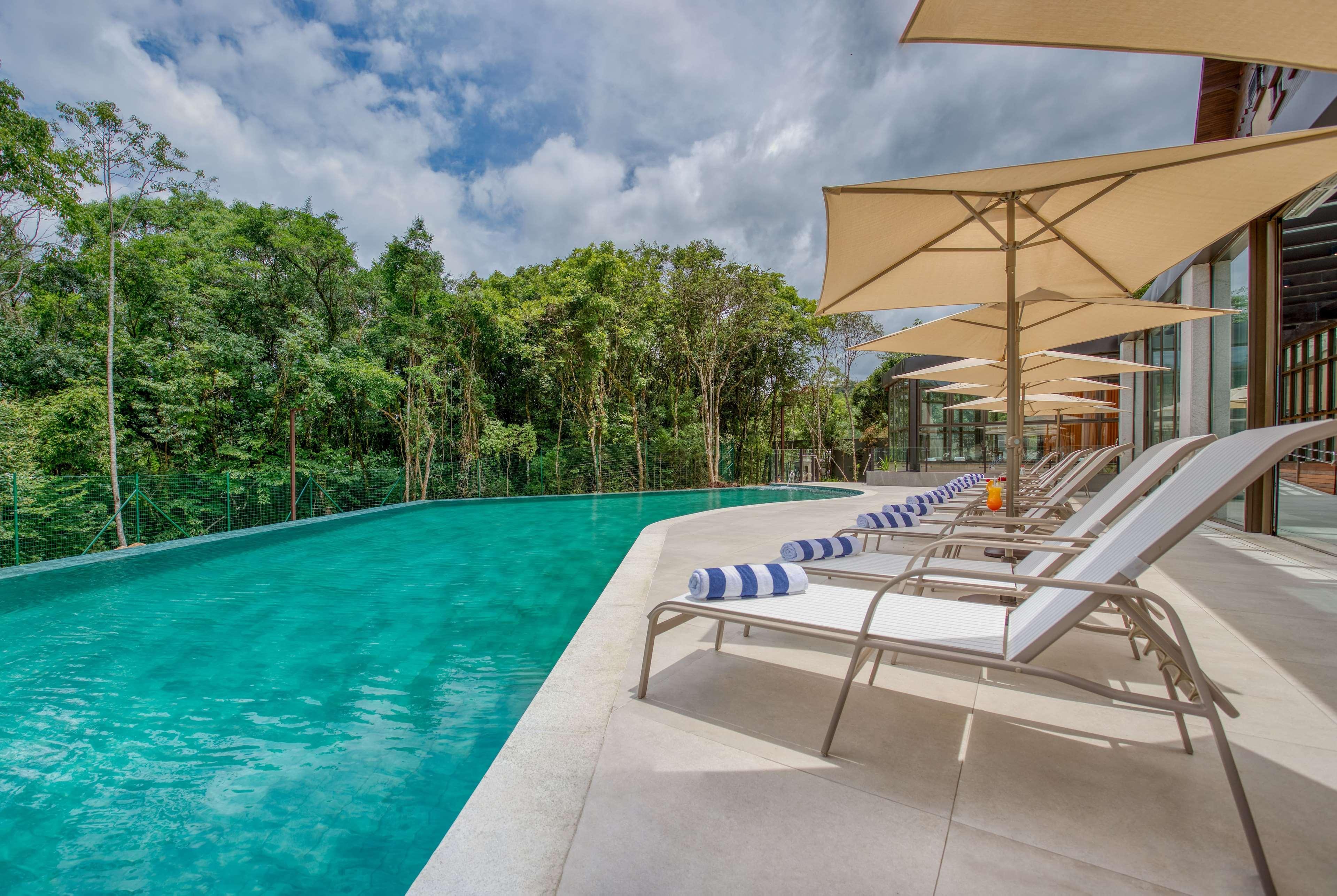 Wyndham Gramado Termas Resort & Spa Exterior foto The photo shows a serene setting featuring a modern infinity pool. The pool has a clear turquoise color and is surrounded by a polished stone deck. Several lounge chairs with white fabric and blue striped cushions are lined up along one side, each wi
