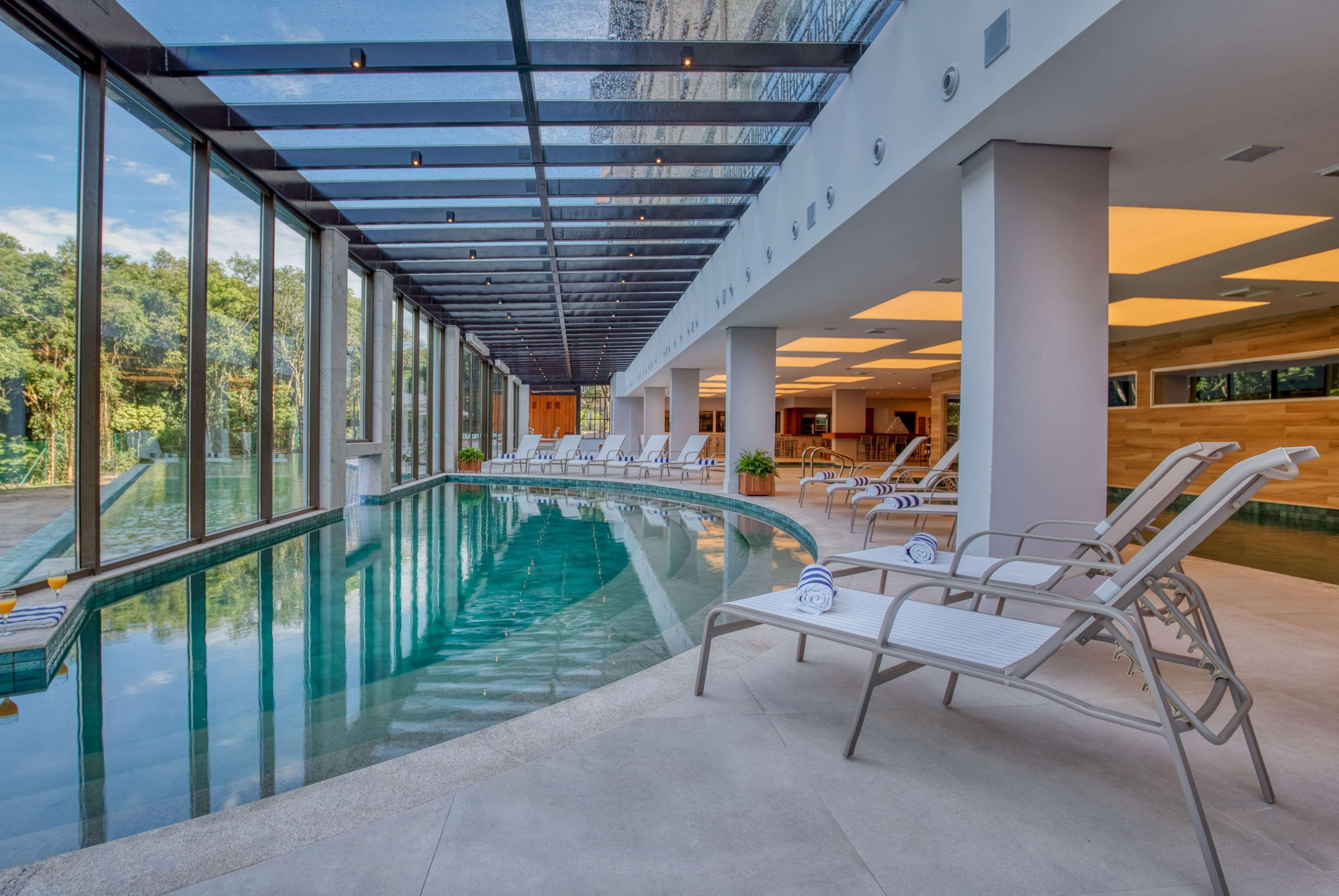 Wyndham Gramado Termas Resort & Spa Exterior foto The photo shows a modern indoor swimming pool area. The pool is surrounded by large glass windows that allow natural light to enter, creating a bright and open atmosphere. There are several white lounge chairs positioned beside the pool, offering a r