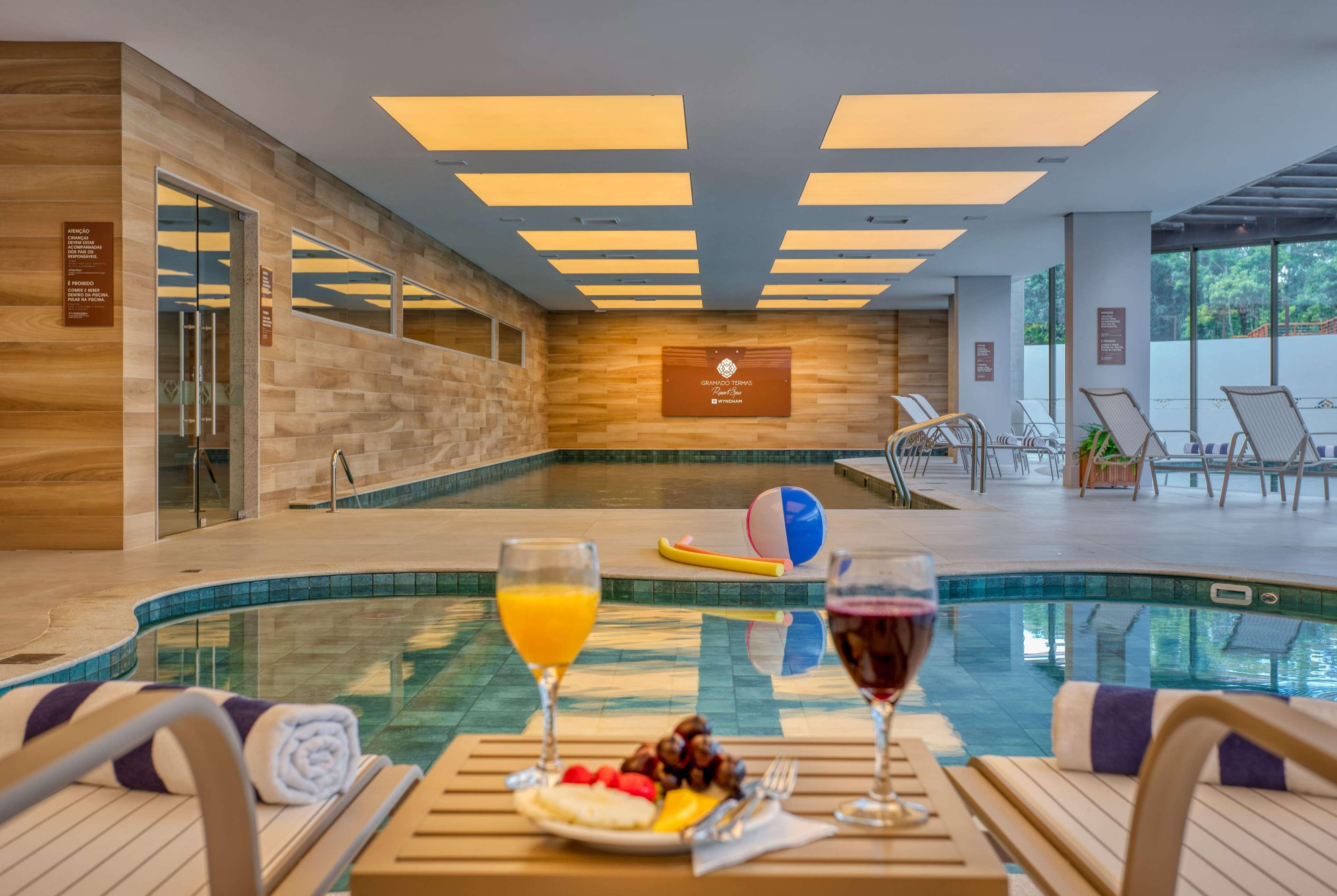 Wyndham Gramado Termas Resort & Spa Exterior foto The image shows an inviting indoor pool area. There are two drinks on small tables, one orange and one red, accompanied by a plate of fruit. The pool is surrounded by lounge chairs and features a smooth, tiled surface. Soft lighting from the ceiling 