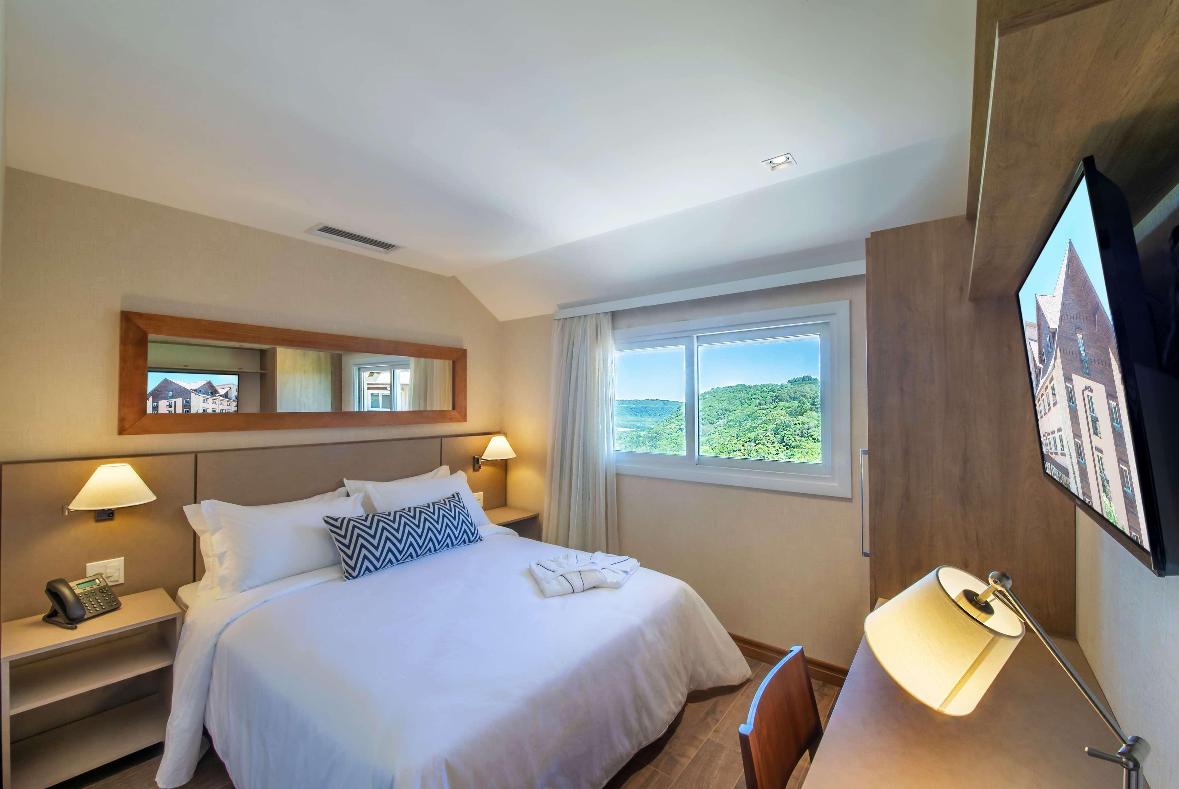 Wyndham Gramado Termas Resort & Spa Exterior foto The photo shows a cozy hotel room featuring a queen-size bed with white linens and a decorative pillow. On either side of the bed are bedside lamps. There is a desk with a modern desk lamp and a flat-screen television mounted on the wall. A window le