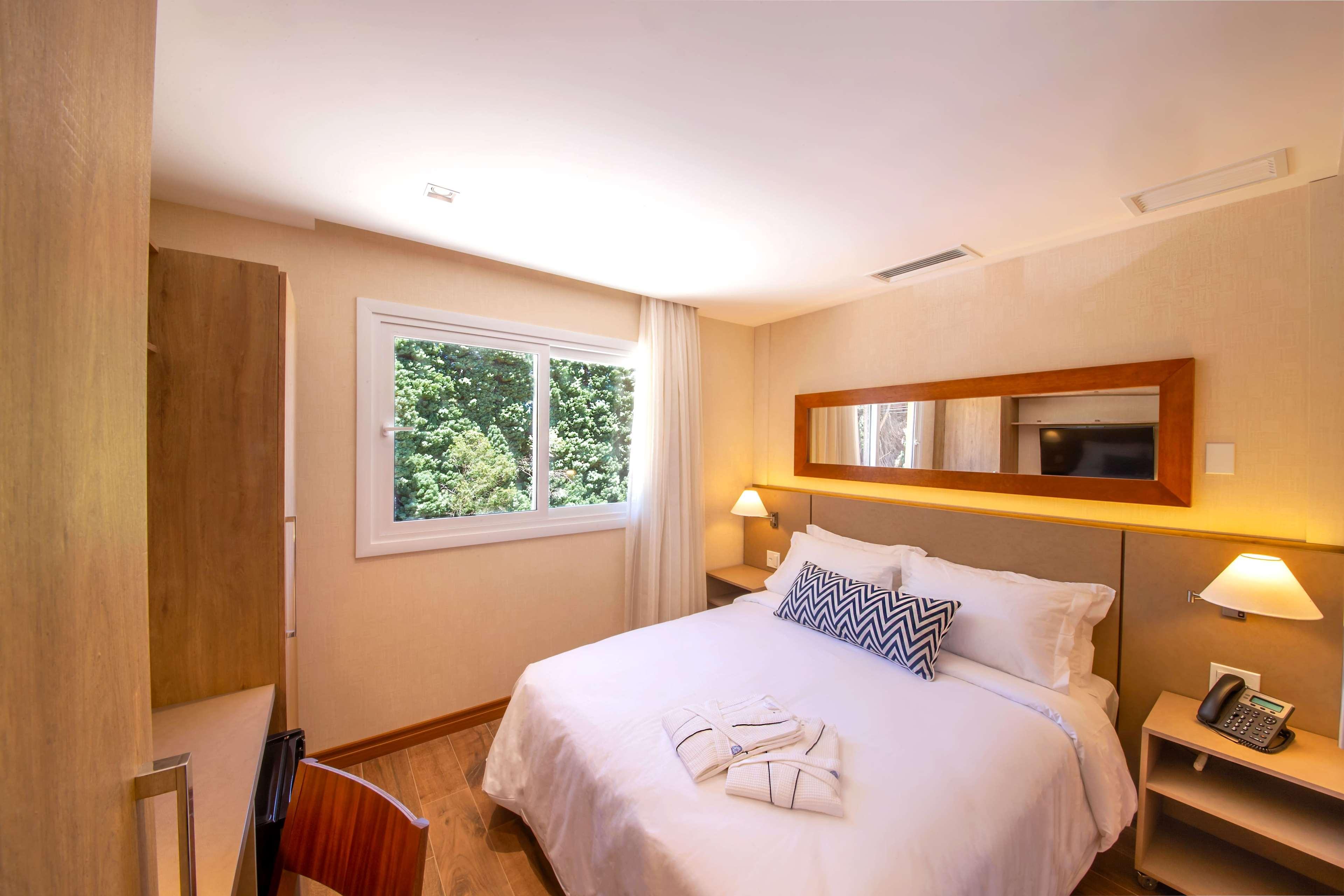 Wyndham Gramado Termas Resort & Spa Exterior foto The photo shows a cozy bedroom featuring a comfortable bed with white bedding and a decorative black-and-white pillow or throw. There is a small desk with a wooden chair, and the room includes a window that offers a view of greenery outside. A mirror