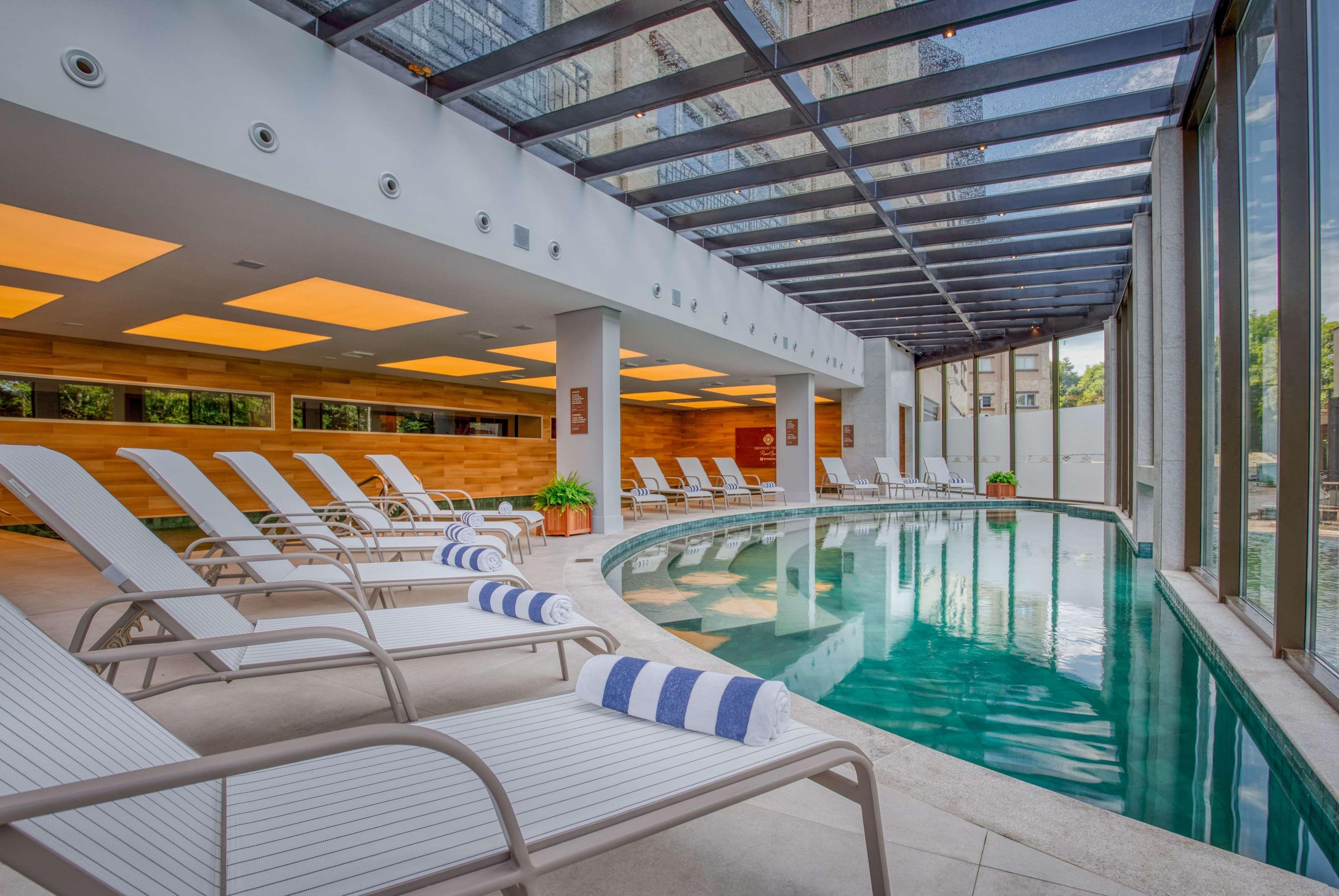 Wyndham Gramado Termas Resort & Spa Exterior foto The photo shows an indoor swimming pool area designed for relaxation. The pool is curved, featuring clear blue water and surrounded by several white lounge chairs. The chairs have blue and white striped towels placed on them. The space is well-lit, w
