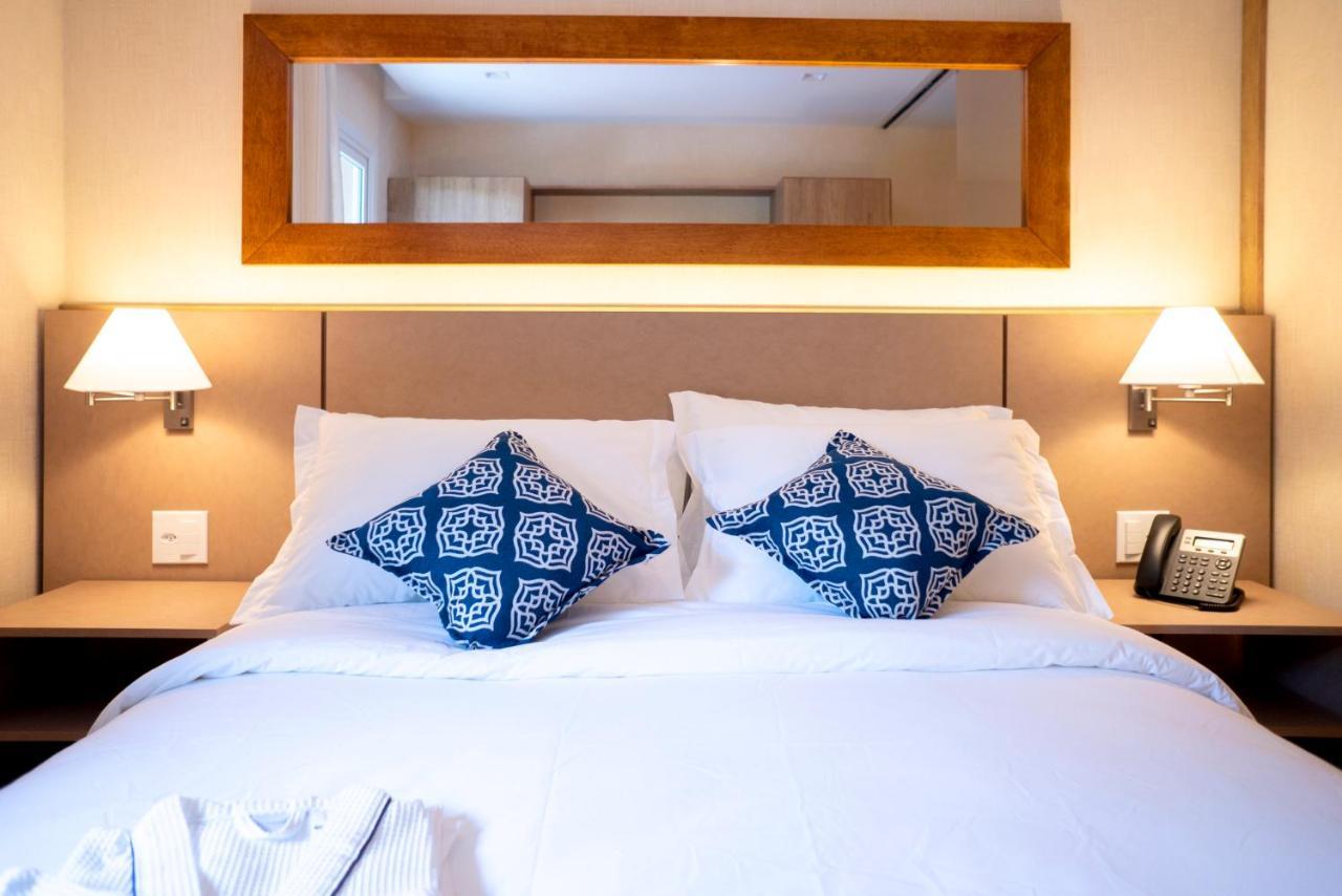 Wyndham Gramado Termas Resort & Spa Exterior foto The photo shows a neatly made bed with white linens and two decorative pillows featuring a blue geometric pattern. Above the bed, there is a wooden-framed mirror reflecting the room's interior. On either side of the bed, there are wall-mounted lamps 